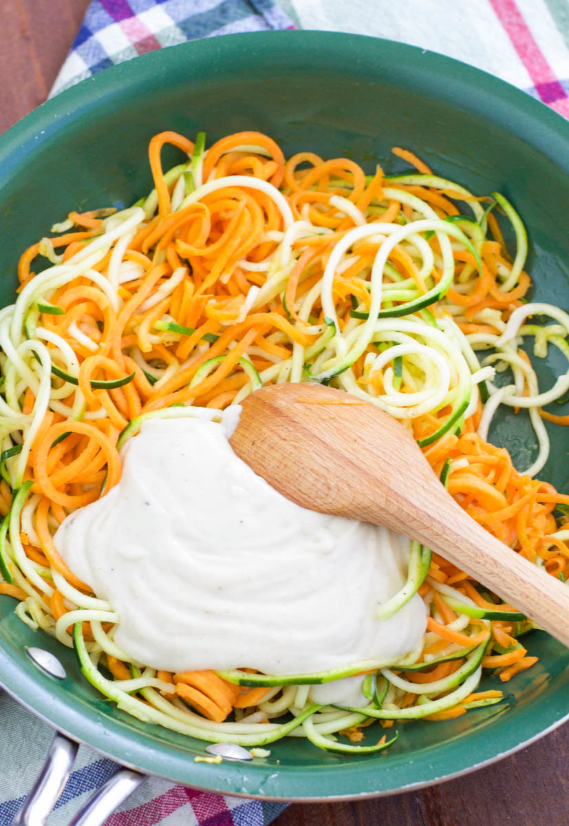 A wooden spoon stirring white sauce into spiraled vegetable noodles in a green pan on top of a plaid napkin. 