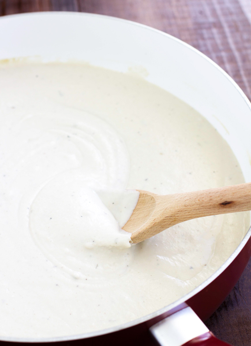 A spoon stirring vegan white sauce in a large pan on a rustic background.