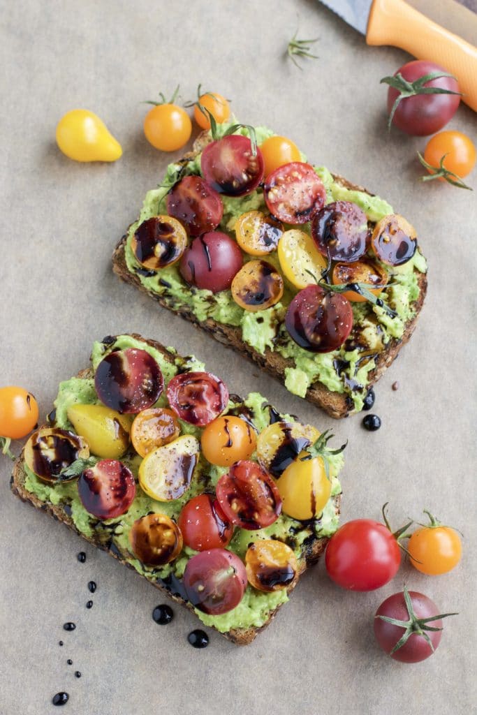 Two slices of avocado toast with tomatoes on top of a piece of parchment paper. 