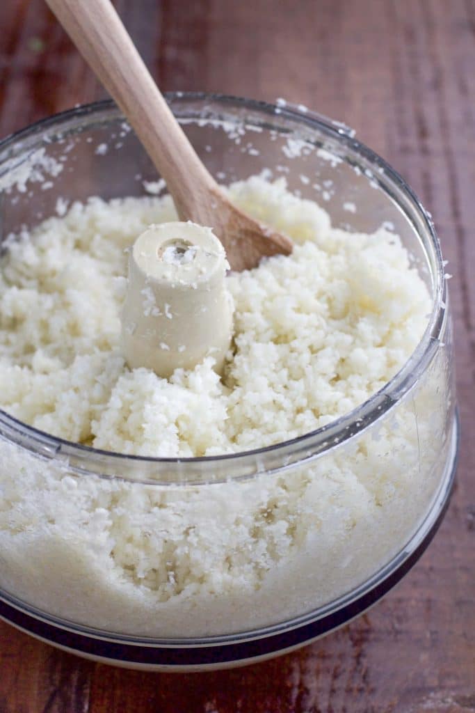 A food processor cup filled with cauliflower rice and a wooden spoon on a rustic table.