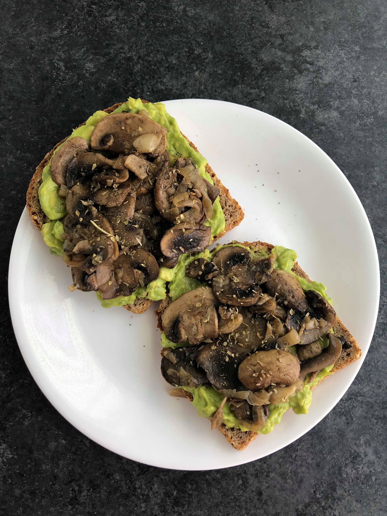 Two slices of avocado toast with mushrooms on top on a white plate with a dark background.