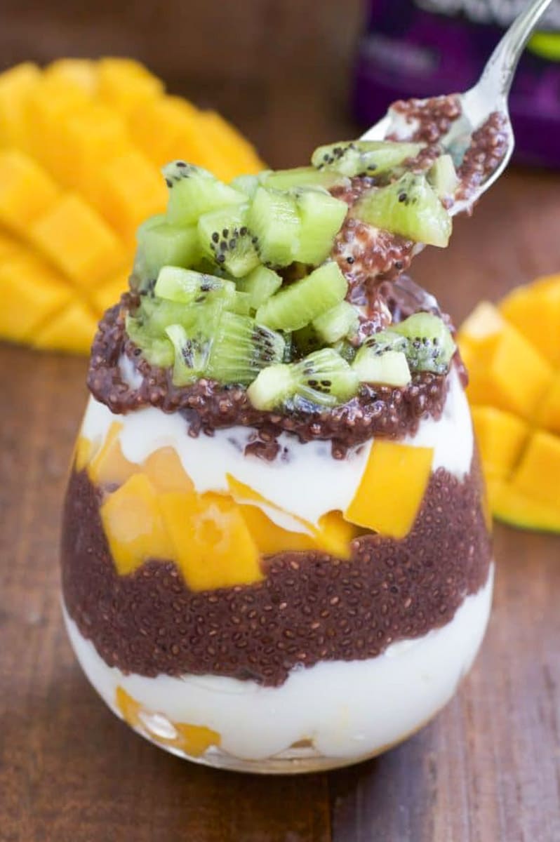 A spoon dipping into a cup filled with layers of yogurt, chia seed pudding, and fresh fruit on a rustic background.