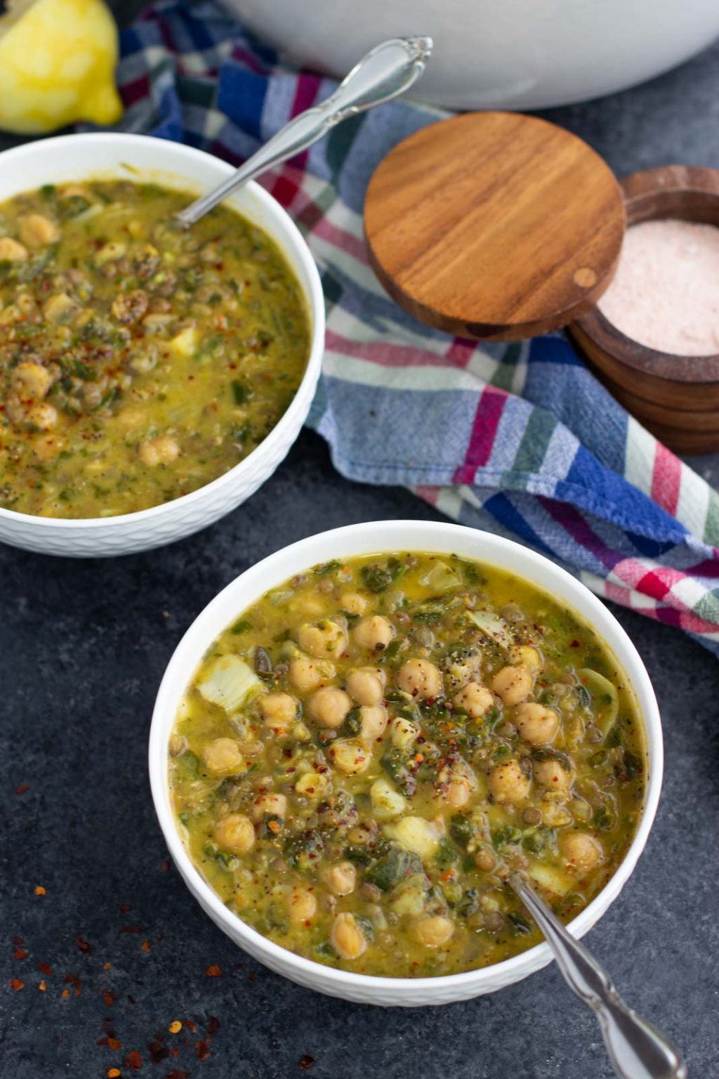 Two white bowls filled with vegan chickpea soup next to a blue plaid towel and a bowl of salt.