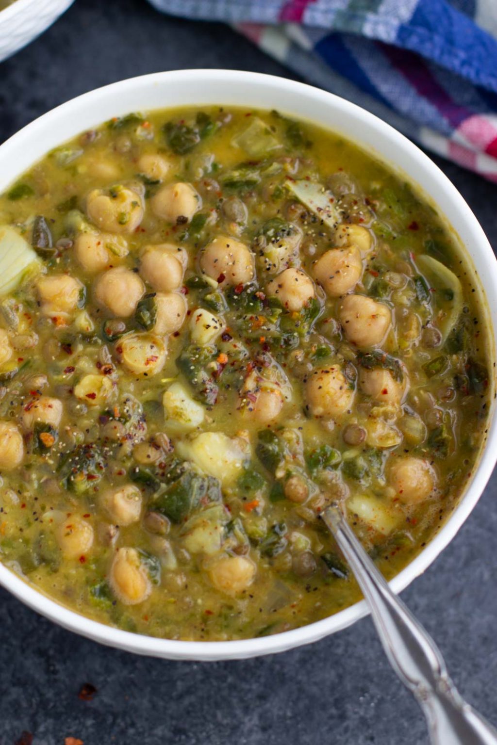 A white bowl filled with vegan chickpea soup and a spoon next to a blue plaid towel.