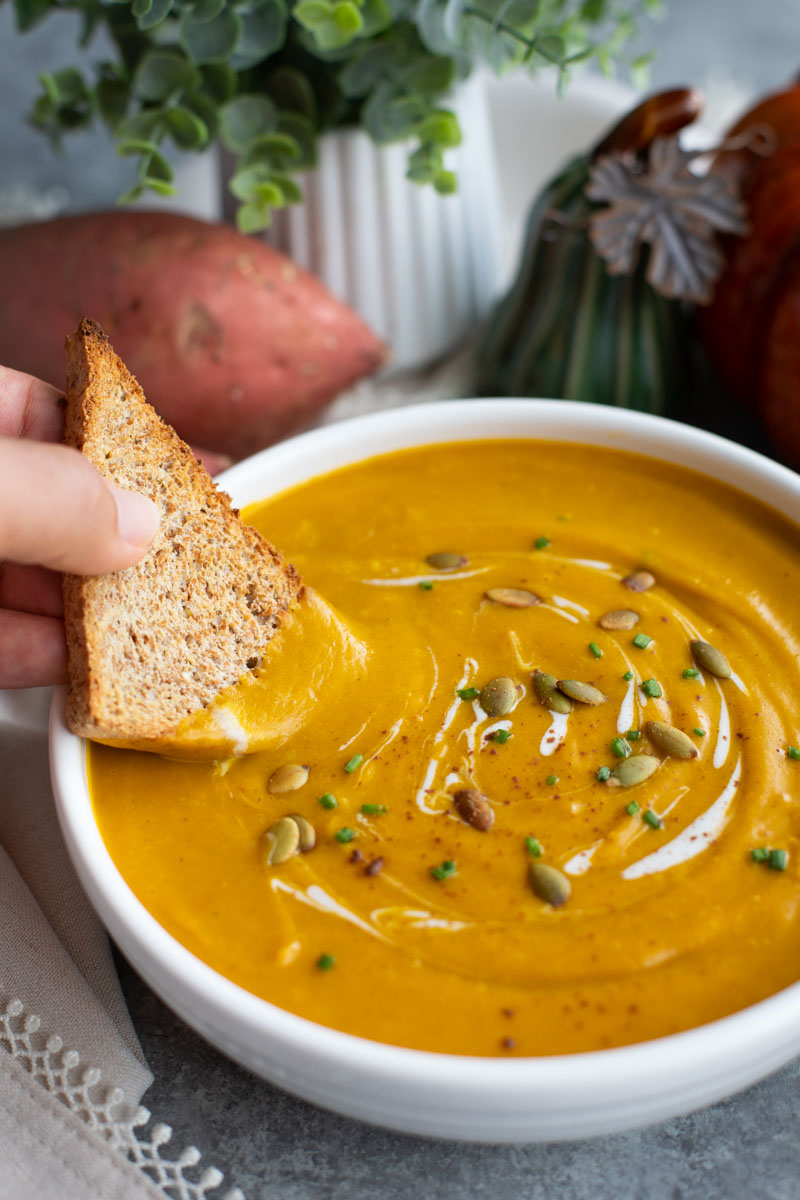 A hand dipping a piece of toasted bread into a bowl of soup next to a sweet potato.