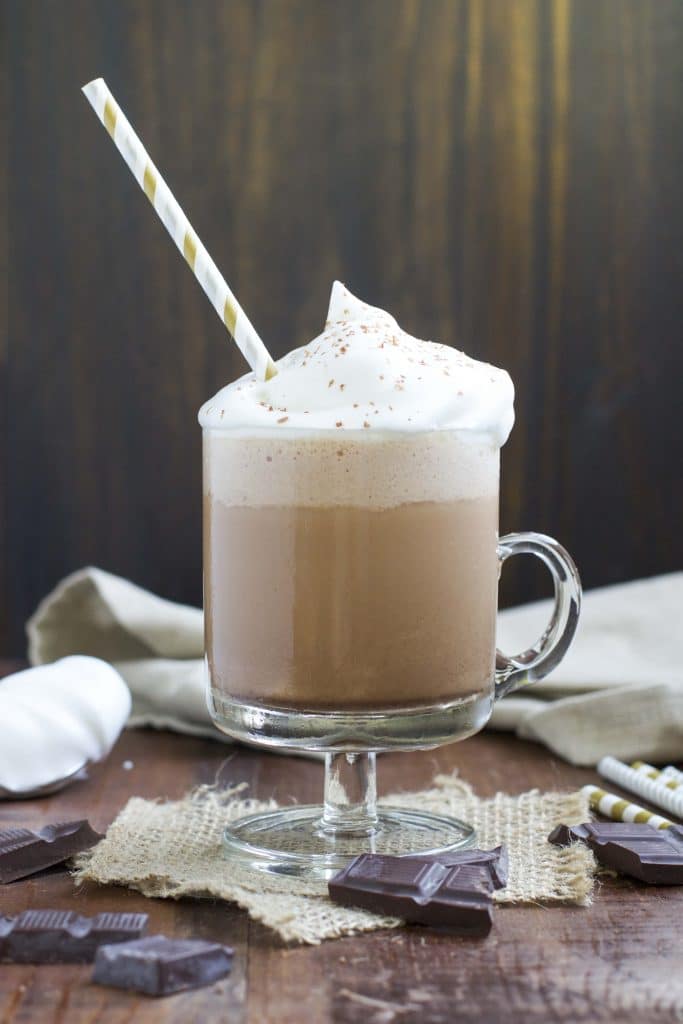 A glass mug filled with a vegan Frappuccino topped with whipped cream next to chunks of chocolate on a rustic background. 