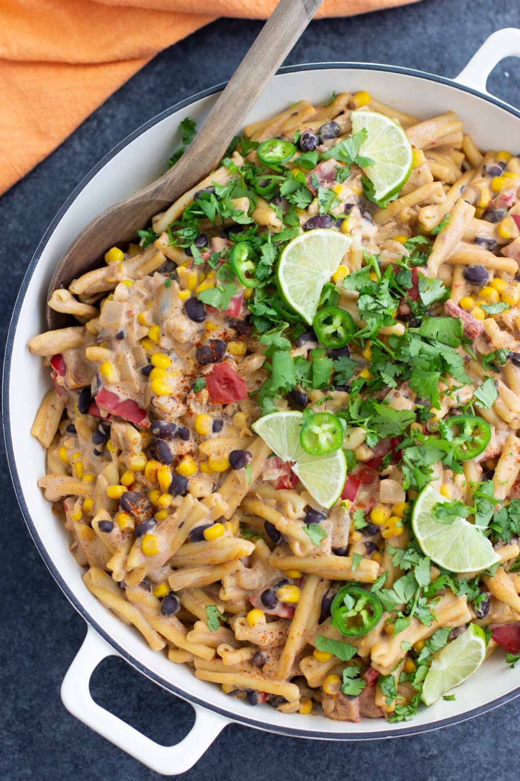 A large white skillet is filled with vegan taco pasta salad and topped with cilantro, lime slices, and jalapeños on a dark textured background. 