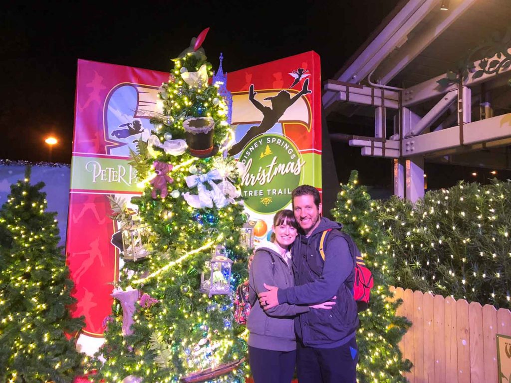 A couple smiling in front of a Christmas tree in the Tree Trail at Disney Springs.