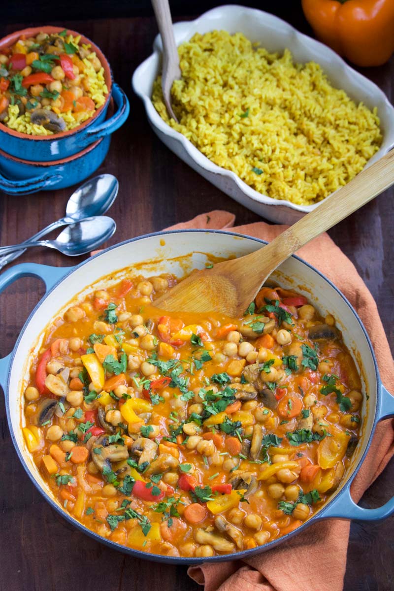 A large bowl of vegetable curry with a wooden spoon next to a few small bowls and a bowl of yellow rice.