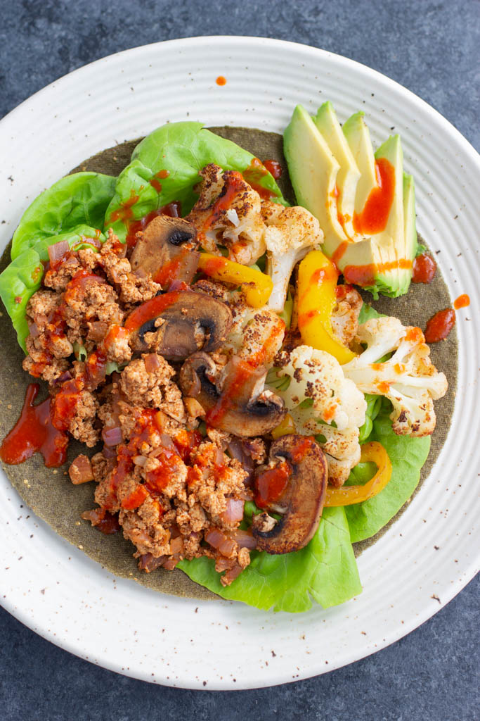 Low-carb vegan tofu tacos filled with roasted vegetables, tofu, and avocado slices in a spinach wrap on a white plate. 