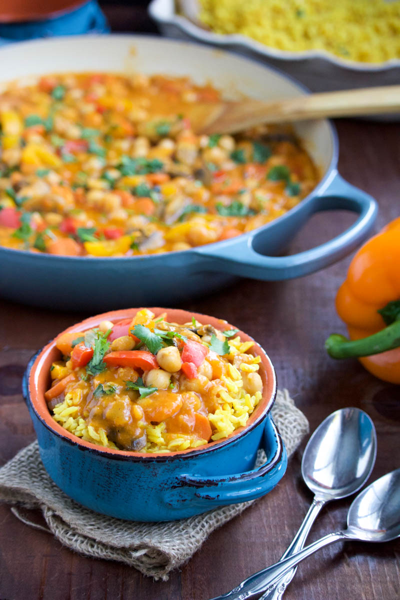 A small blue bowl filled with chickpea and mushroom curry over yellow rice next to a large blue pan of curry. 