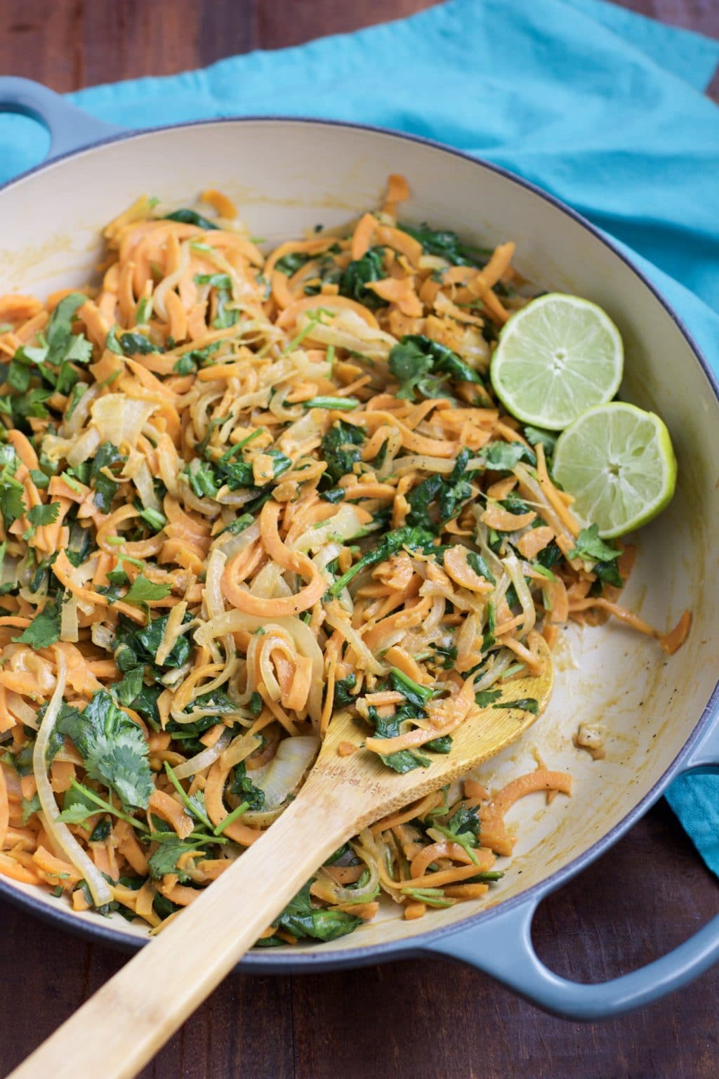 A blue pan filled with spicy sweet potato noodles, a wooden spoon, and sliced limes. 