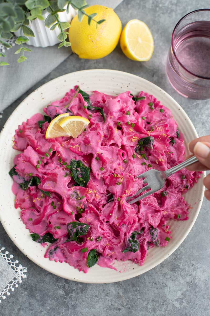 A hand holding a fork over pink noodles on a white plate next to a lemon.