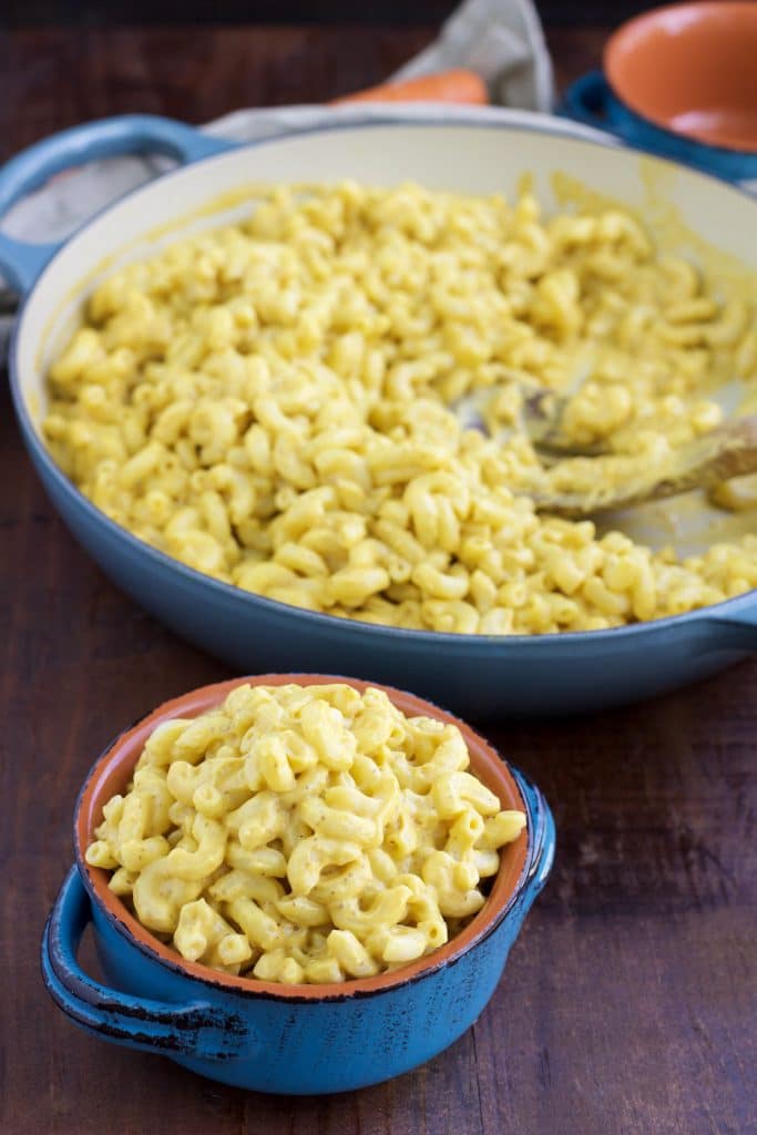 A small blue bowl filled with plant-based Mac and cheese next to a larger pan on a rustic background.
