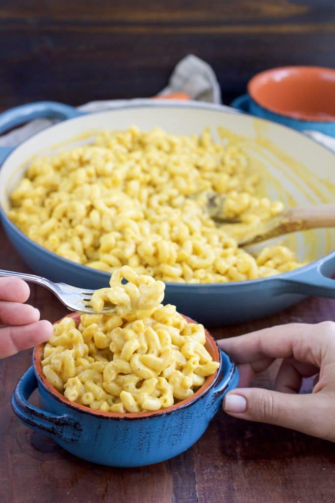 Hands holding a small blue bowl of pasta next to a large pan filled with pasta on a rustic background. 