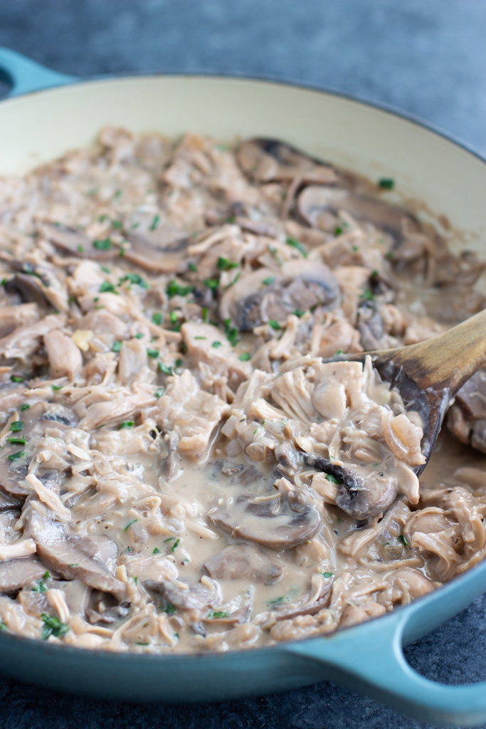 A large blue pan filled with mushroom stroganoff being stirred by a wooden spoon on a dark textured background.