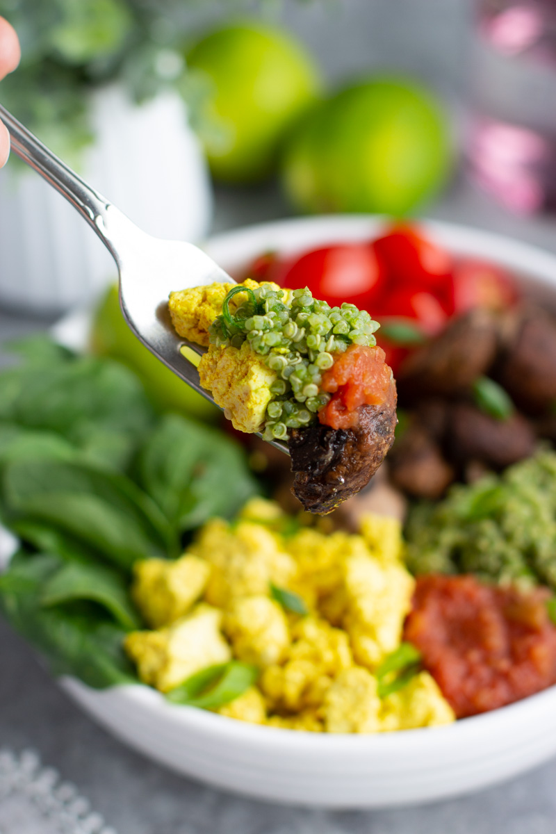 A fork holding quinoa, tofu scramble, and a mushroom over a bowl of vegetables. 