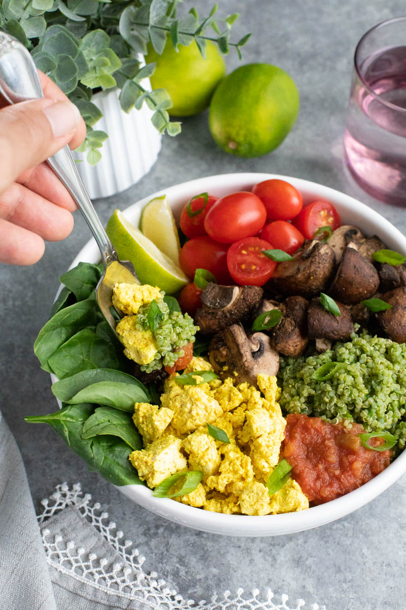 A hand sticking a fork into the spinach in a bowl full of vegetables on a gray background.