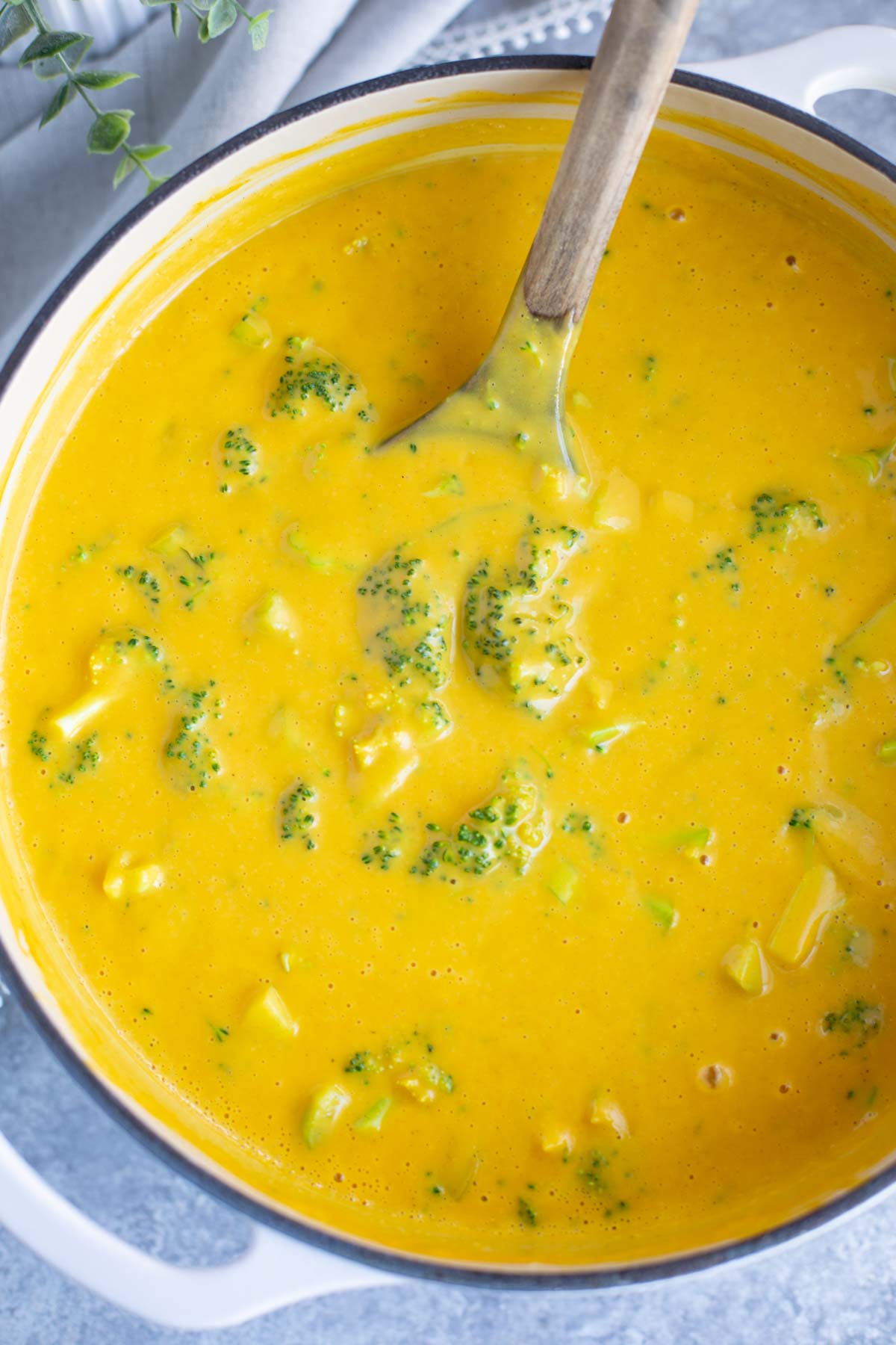 A white dutch oven filled with vegan broccoli cheddar soup and a wooden spoon on a gray background.
