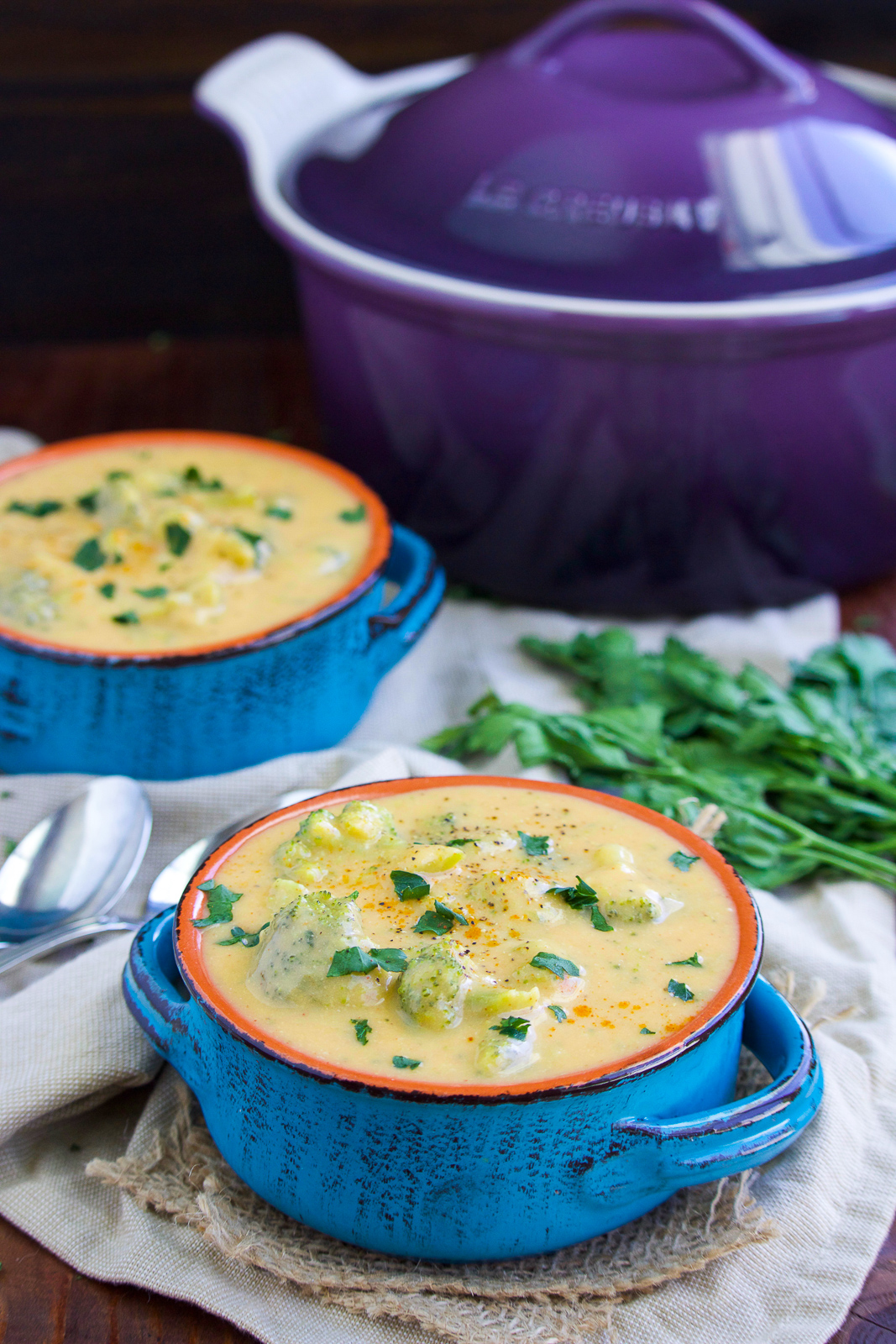 Two blue bowls filled with soup next to fresh parsley and a purple pot on a rustic background.