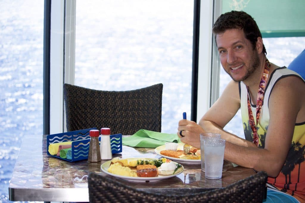 A man smiling and eating lunch at a table by a window overlooking the ocean on a Disney cruise. 