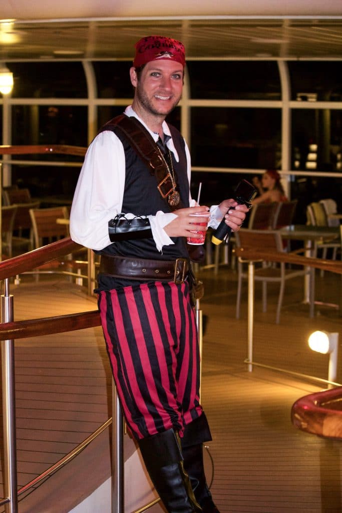 A man dressed in costume as a pirate holding a cup and leaning against a railing on a disney cruise ship. 