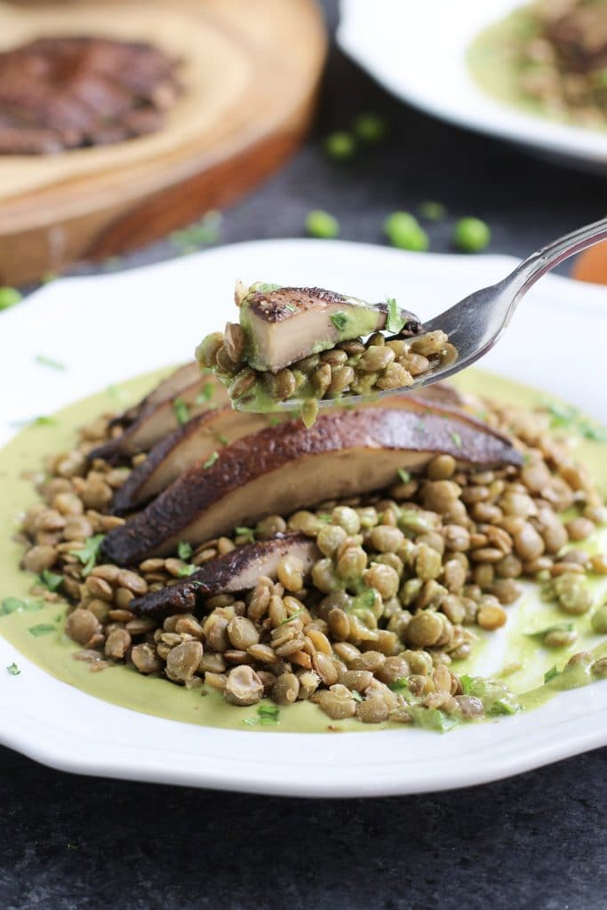 A fork holding a piece of portobello mushroom over a white plate filled with lentils and green sauce.