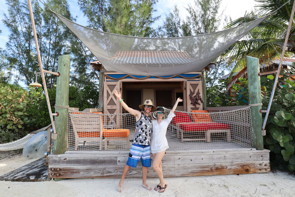 A man and woman posing and smiling with the arms up in front of a Serenity Bay cabana on Castaway Cay. 