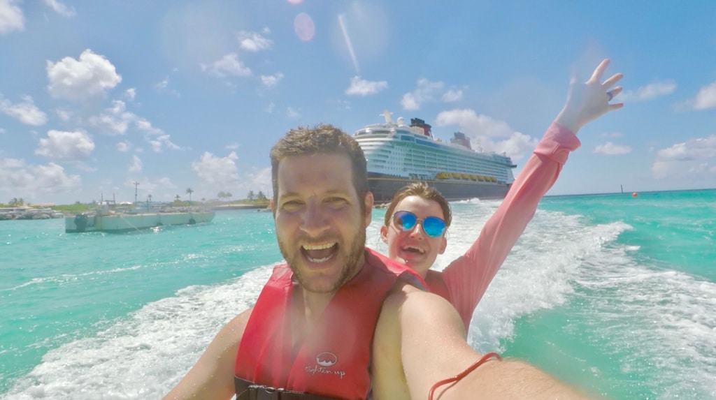 A man and woman riding on a jet ski excursion at Castaway Cay with a Disney cruise behind them. 