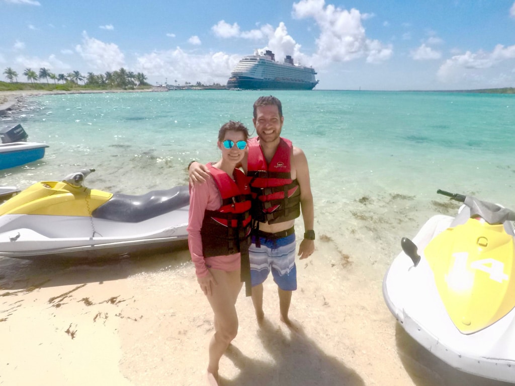 A happy couple standing on the beach next to jet skis in front of the Disney Fantasy on Castaway Cay.