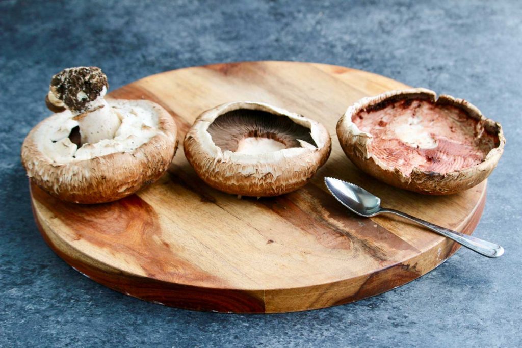 Three portobello mushrooms in varying stages of being cleaned on top of a cutting board.