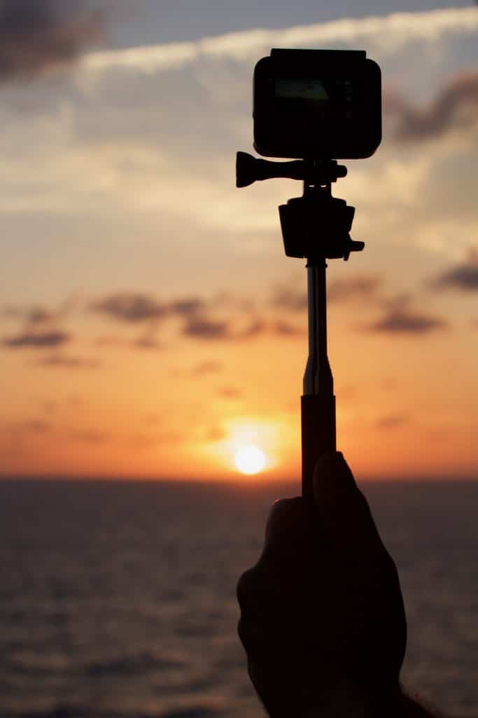 A hand holding a GoPro camera in front of a sunset on the water. 