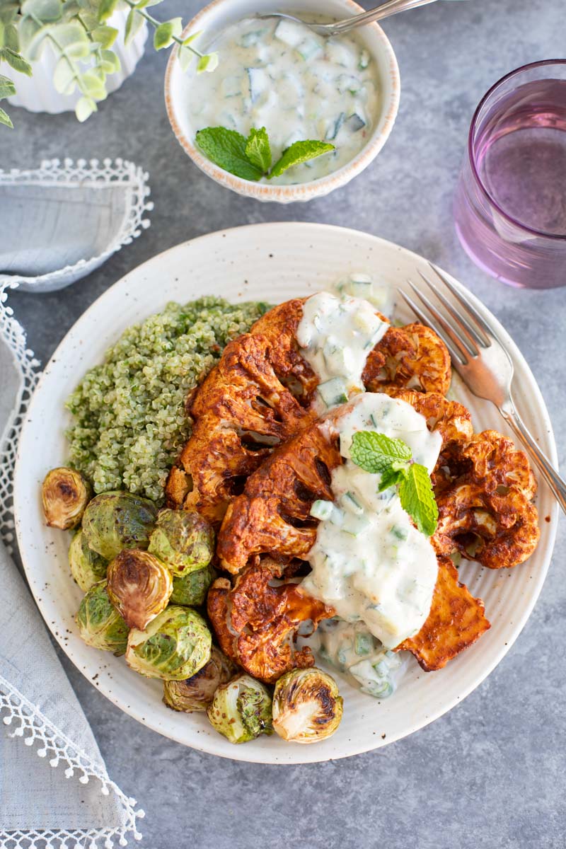 A plate filled with cauliflower steak, Brussels sprouts, and quinoa next to a small bowl of white sauce on a gray background.