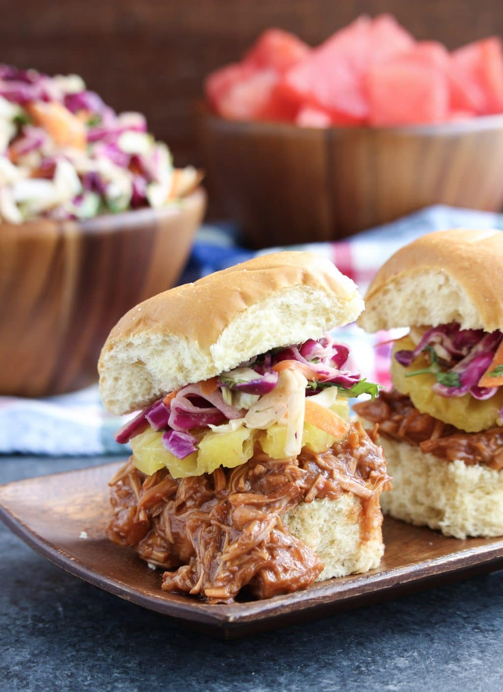 Two sliders on a wooden tray next to bowls of coleslaw and watermelon.