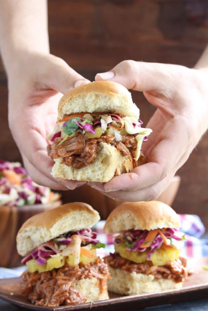 Two hands holding a vegan pulled pork slider over a tray of more sliders on a rustic background. 
