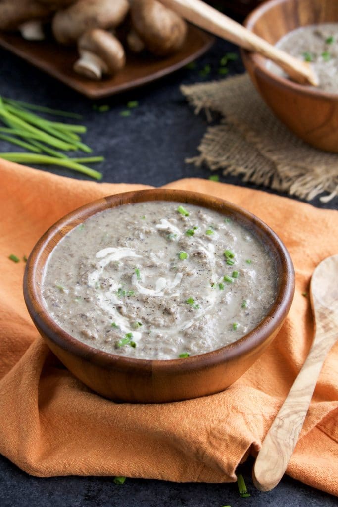 A wooden bowl of low-carb vegan mushroom soup sitting on top of an orange napkin with mushrooms and another bowl of soup in the background.