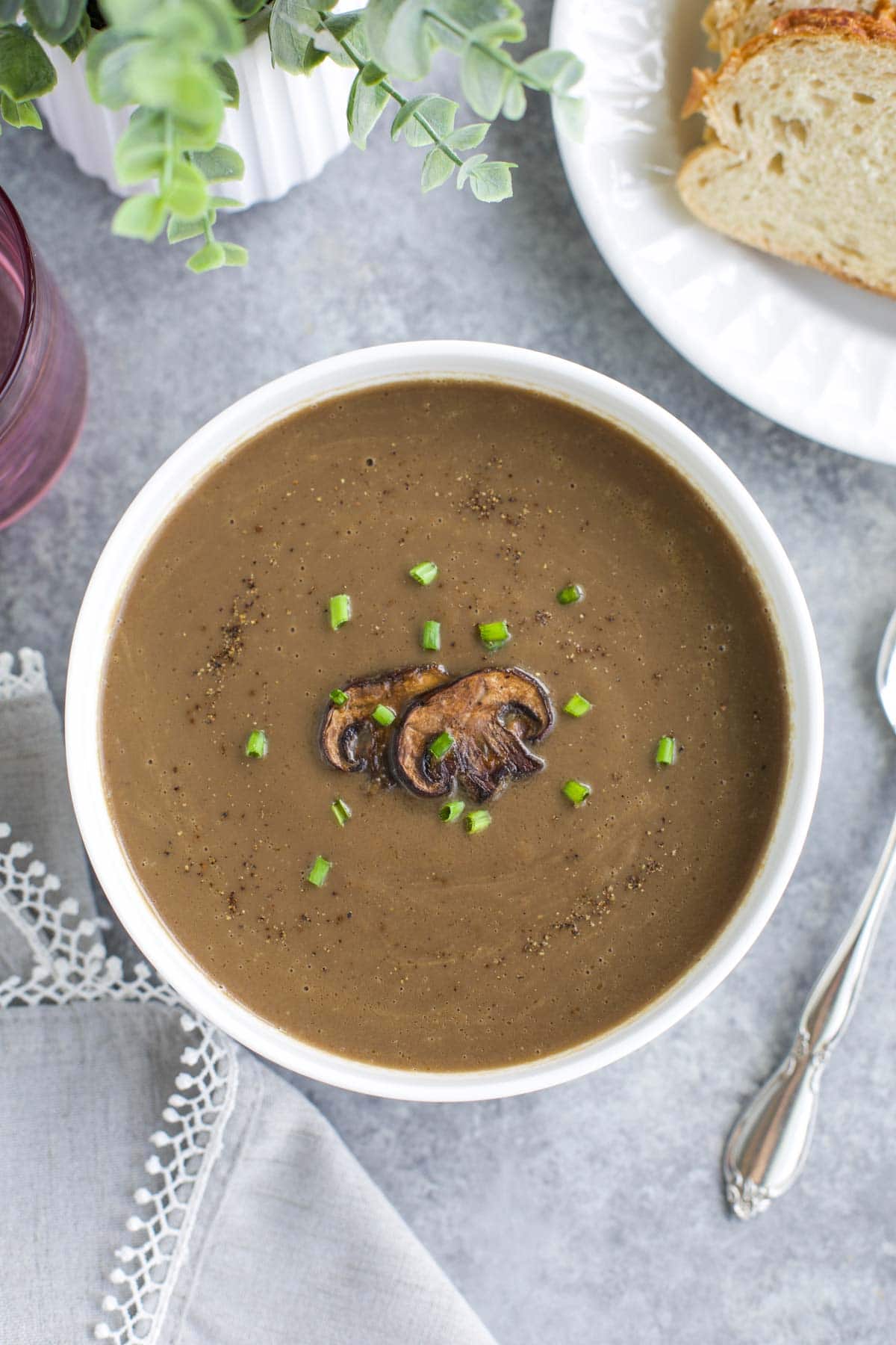 Overhead shot of a white bowl filled with vegan mushroom soup next to a plate of bread on a gray background. 