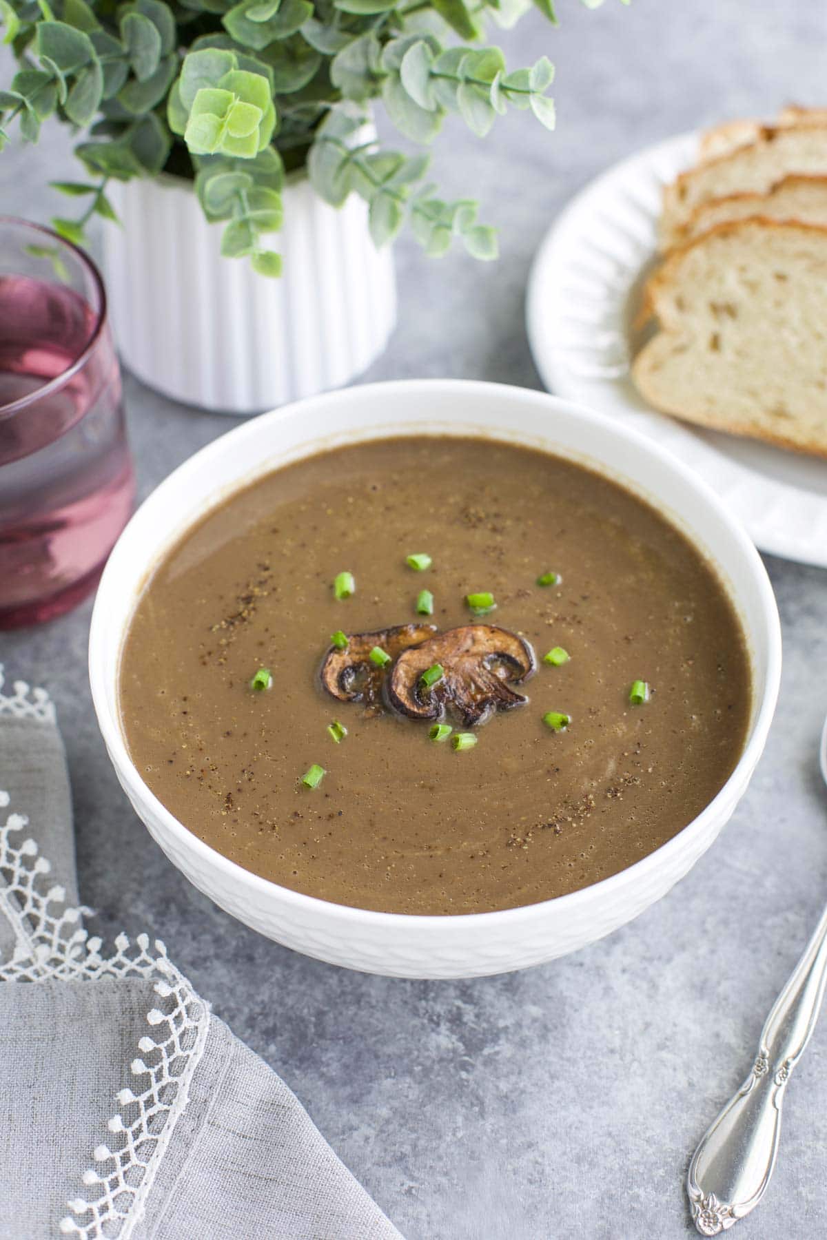 A white bowl filled with soup next to bread slices and a plant on a gray background. 