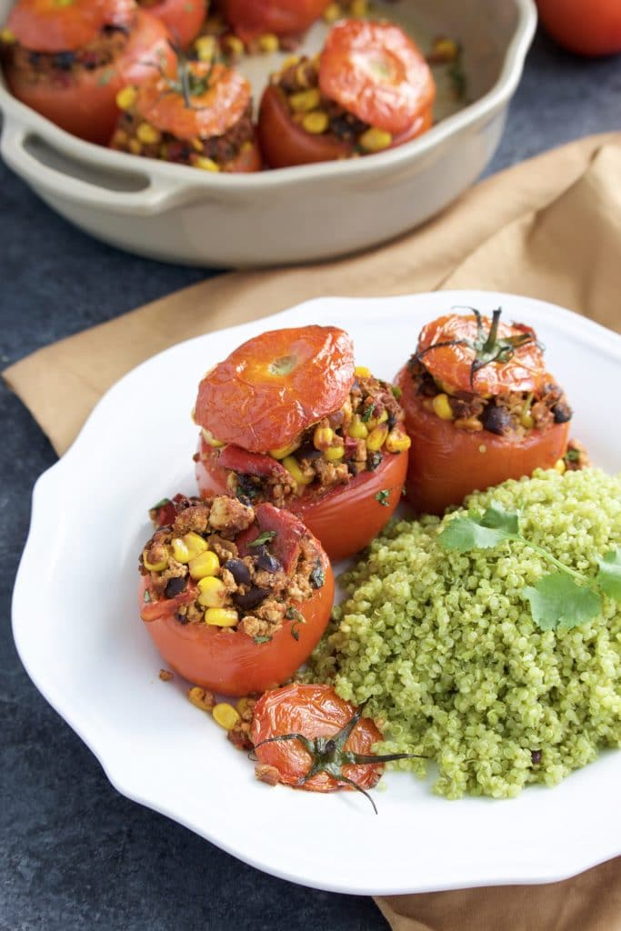 Stuffed tomatoes and cilantro quinoa on a white plate next to a brown napkin and a casserole dish of stuffed tomatoes. 