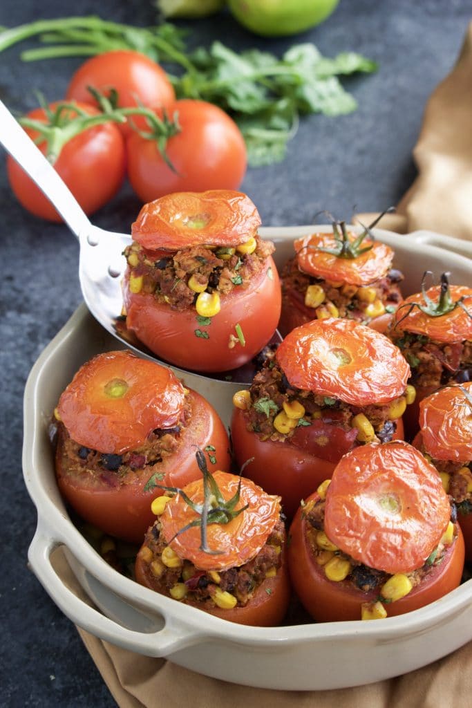 A spoon holding a stuffed tomato over a filled casserole dish on a dark background. 