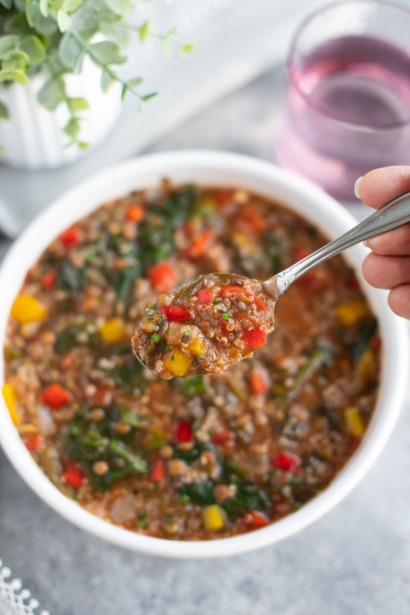 A hand holding a spoonful of soup over a white bowl of soup on a gray background.