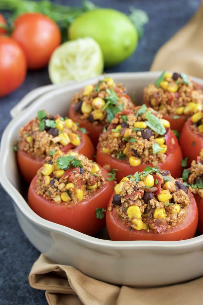 A casserole dish filled with stuffed tomatoes on top of a brown napkin that's next to vegetables. 