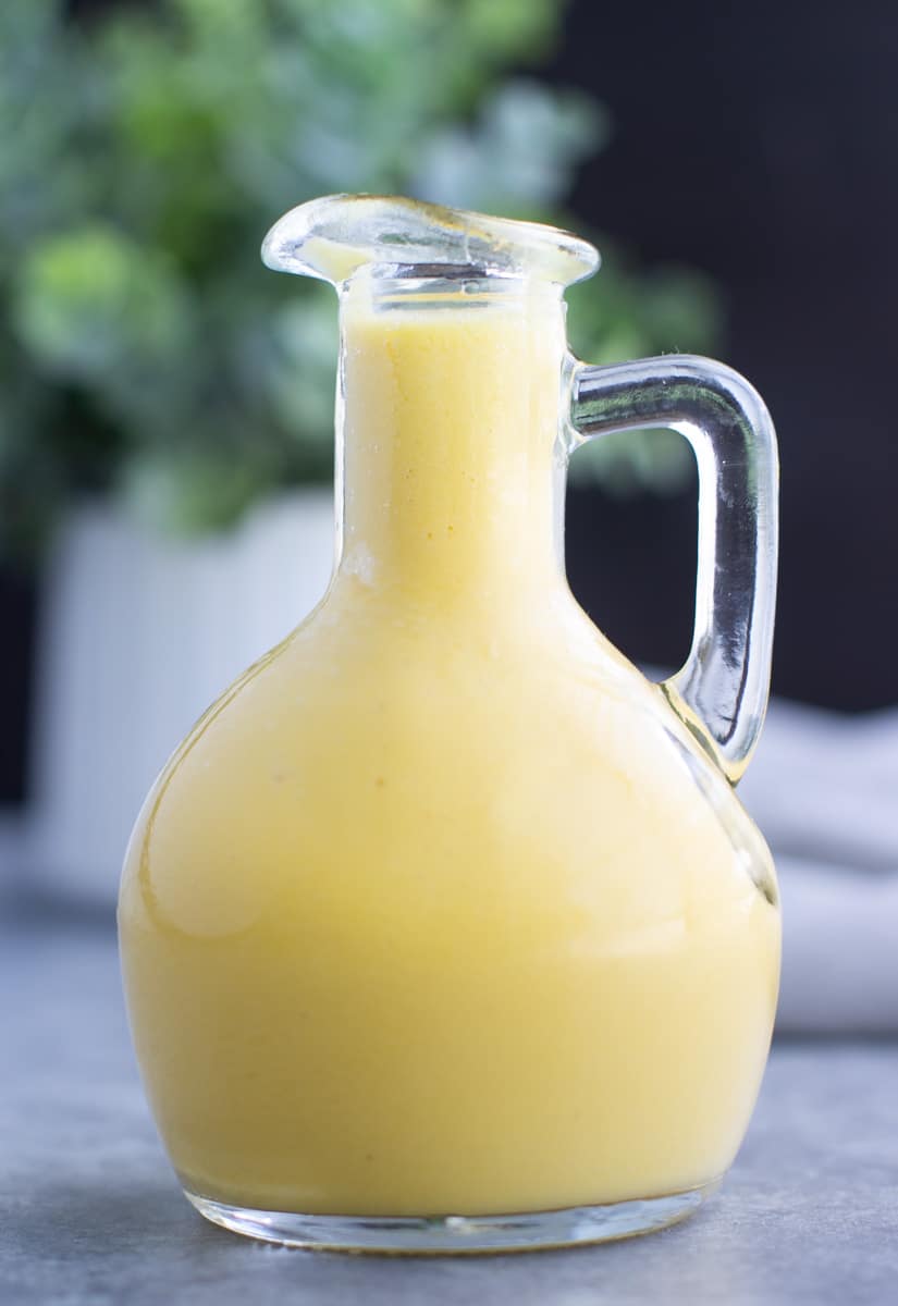 A small glass container filled with yellow mango salad dressing in front of a small plant. 