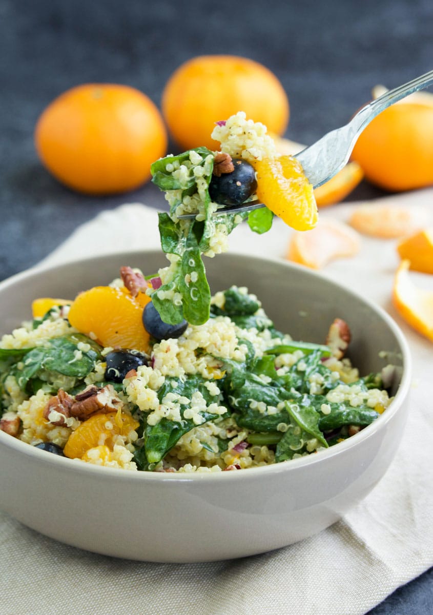 A fork being dipped into a bowl of quinoa salad on top of a napkin next to a couple of oranges.