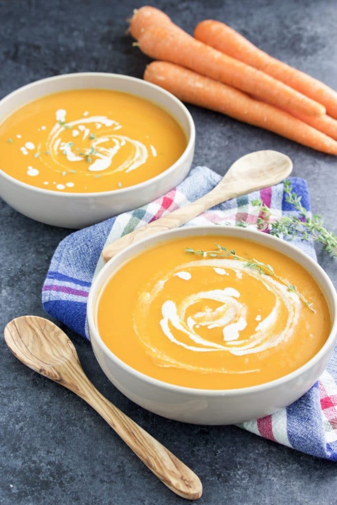 Two bowls of soup next to a plaid napkin, wooden spoons, and a few carrots on a dark background. 