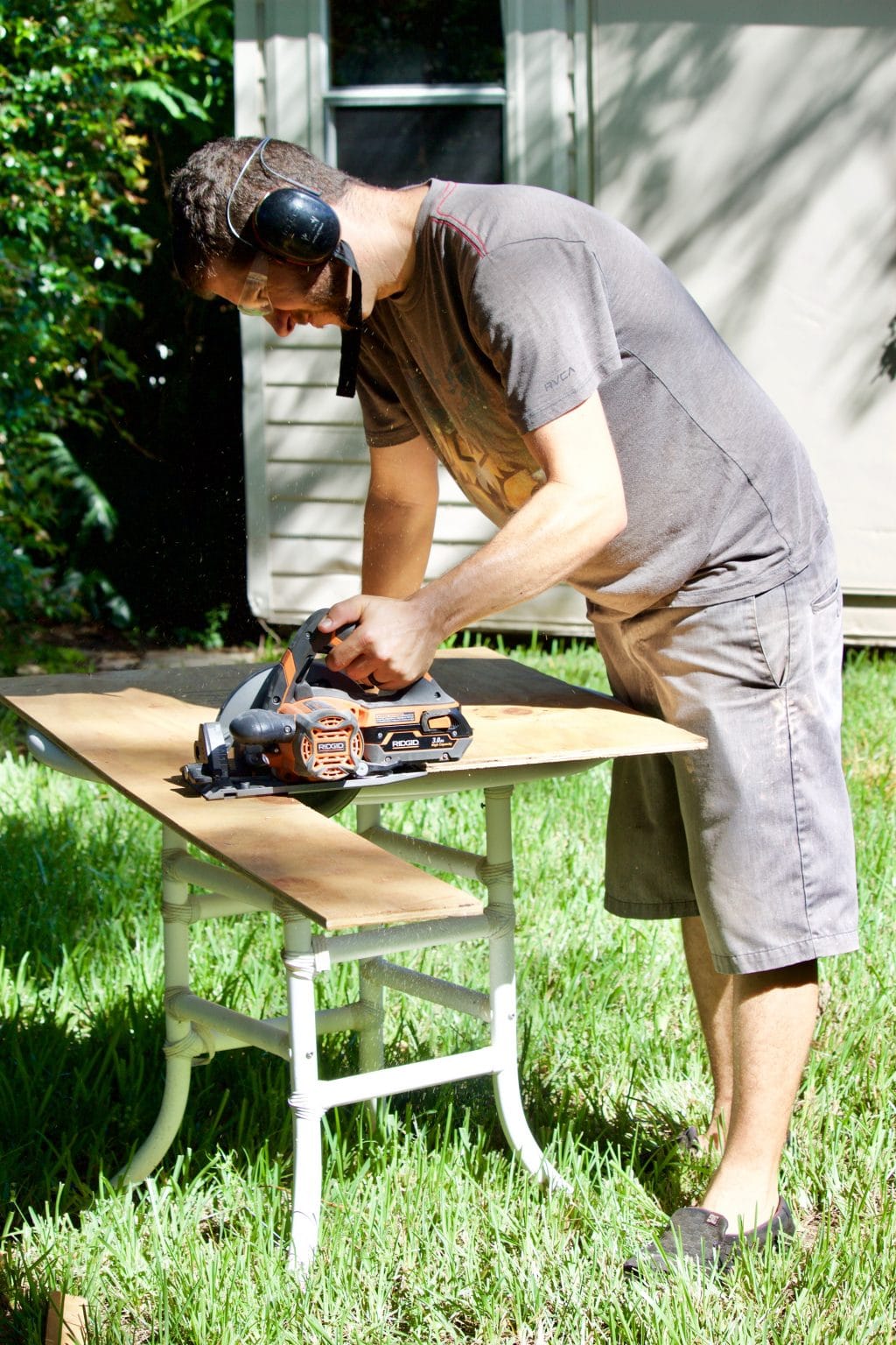 A man with a table saw cutting a piece of plywood on a table over grass. 