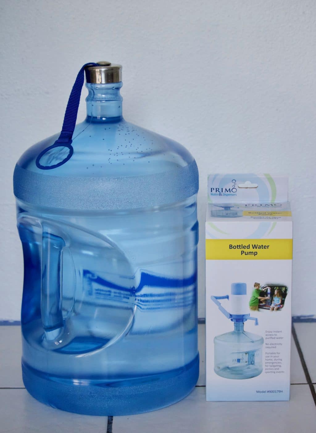 A large 5-gallon water bottle next to a hand pump on a white tile floor. 