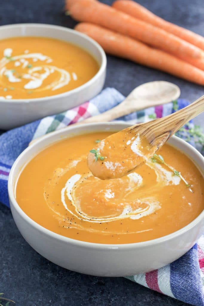 A wooden spoon dipping into a bowl of vegan carrot ginger soup next to another bowl of soup and a plaid napkin on a dark background.