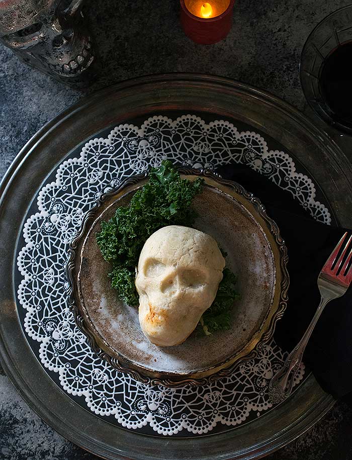 An edible skull on top of a dark plate and matching charger with a fork and a candle. 