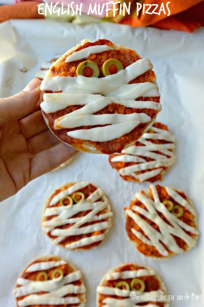 A hand holding a vegan English muffin mummy pizza for Halloween over a white background.